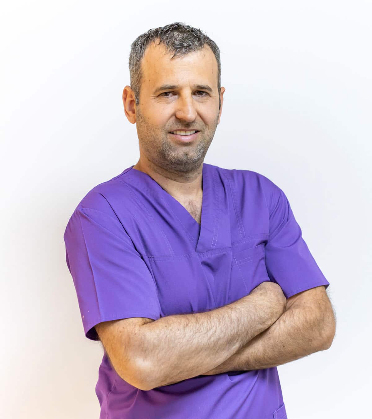 O persoană cu păr scurt și miriște, purtând un top de scrub violet, stă cu brațele încrucișate pe un fundal simplu.