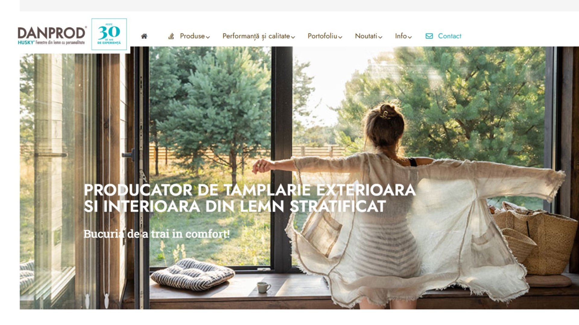O femeie stă cu brațele întinse în fața unor ferestre mari de sticlă, cu vedere la un peisaj exterior verde. Textul în limba română promovează produsele de tâmplărie din lemn.