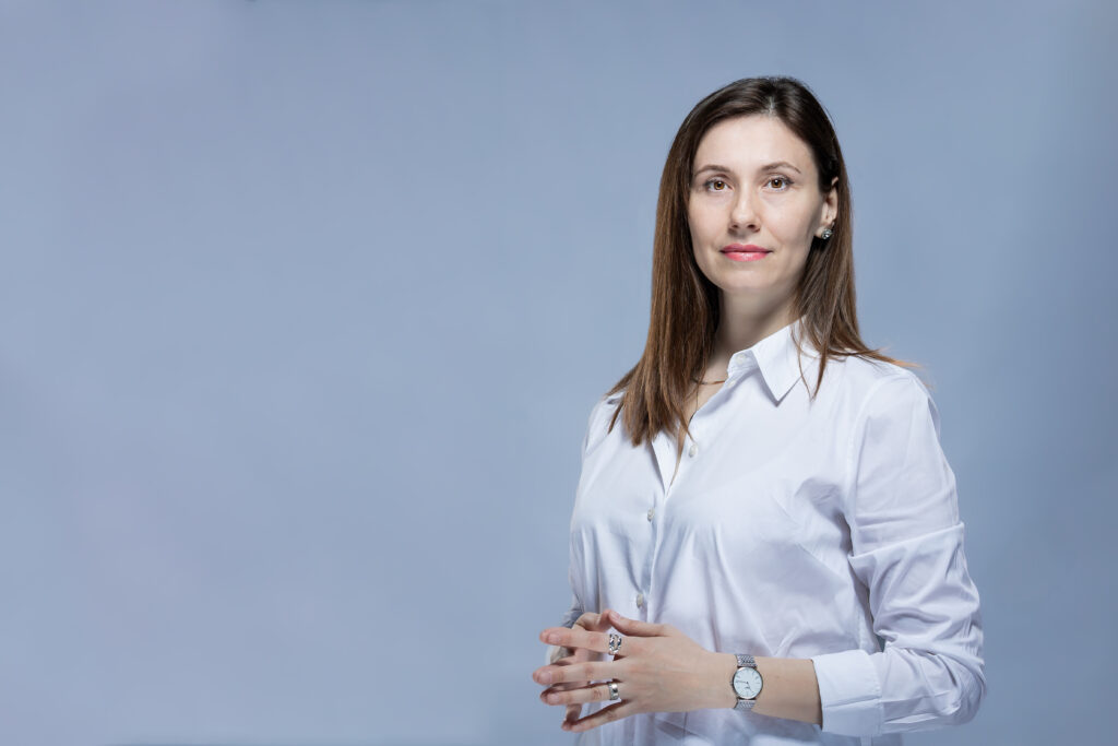 Femeie cu păr șaten drept, purtând o cămașă albă cu nasturi și ceas, în picioare pe un fundal albastru deschis, privind spre camera foto - perfect pentru a reprezenta Servicii SEO București cu o imagine profesională și accesibilă.
