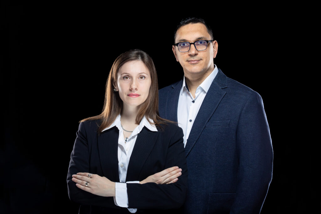 Două persoane în costume de afaceri stau împreună pe un fundal negru, reprezentând Servicii SEO București. Femeia are brațele încrucișate, iar bărbatul stă puțin în spatele ei. Ambii au expresii neutre.