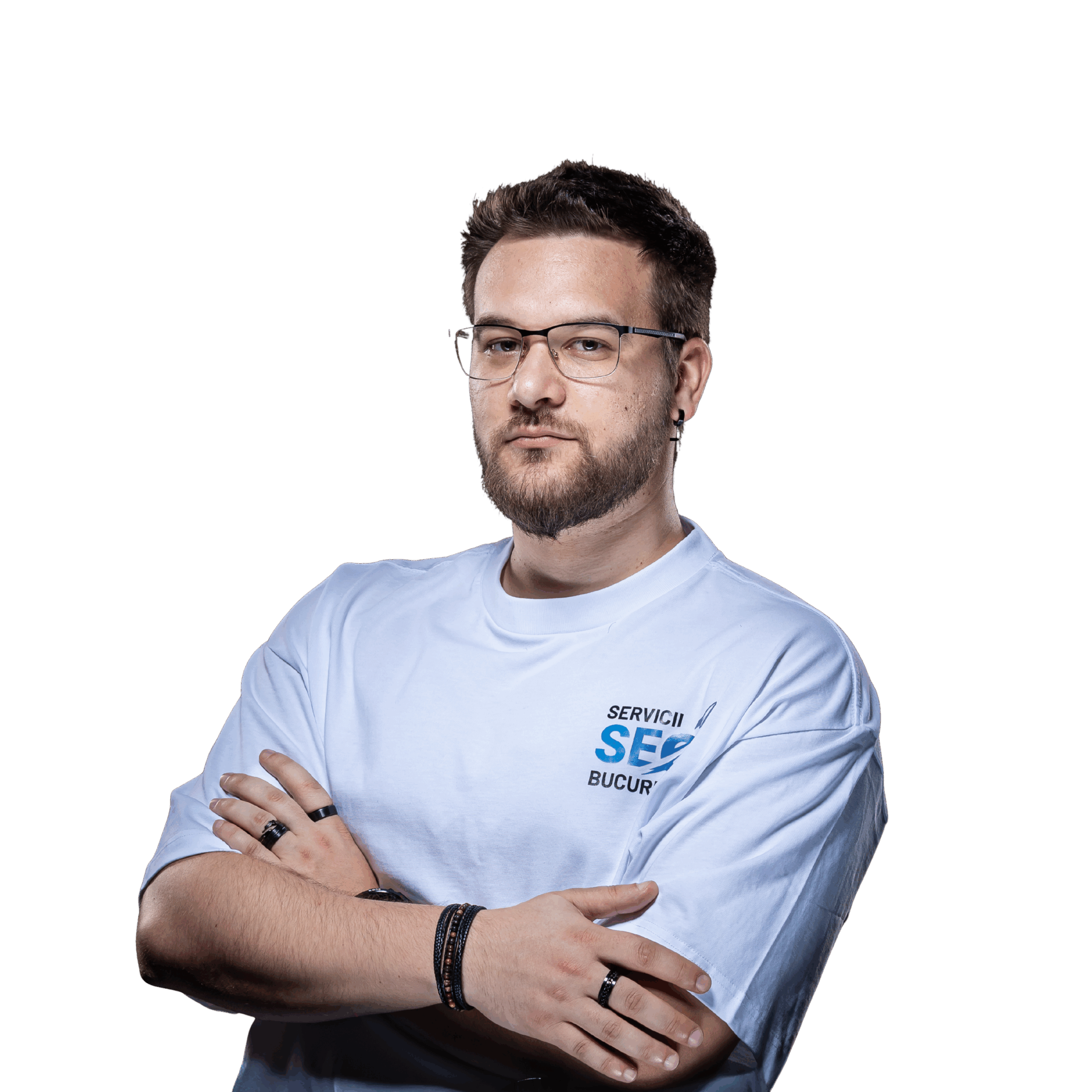 Un bărbat cu părul scurt, ochelari și barbă stă cu brațele încrucișate, purtând un tricou albastru deschis și brățări, pe un fundal alb simplu - emană încredere ca un expert SEO gata să facă față provocărilor digitale.
